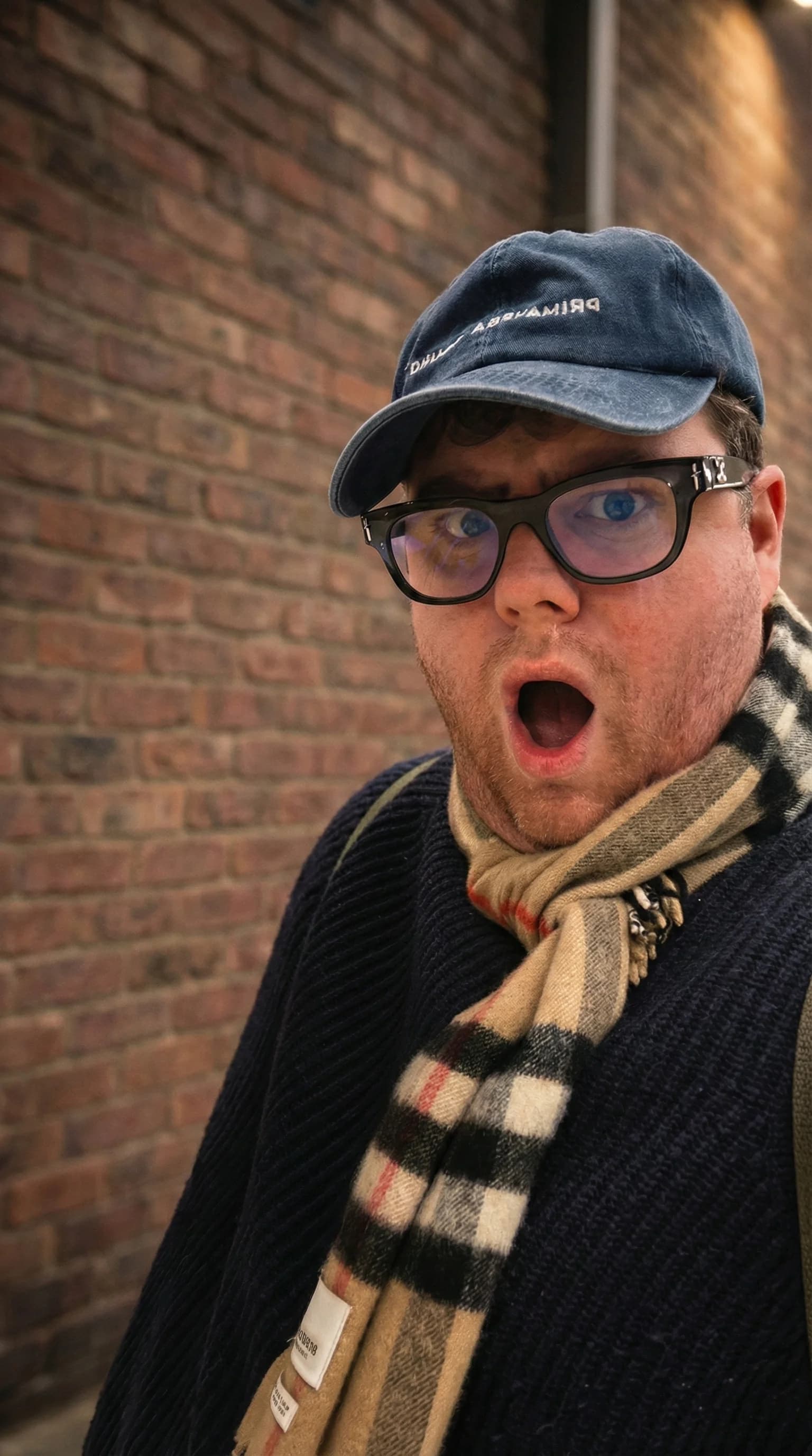 Photograph of James Rooney in a navy cap, glasses and a check scarf, mouth open in mock surprise, against a brick wall